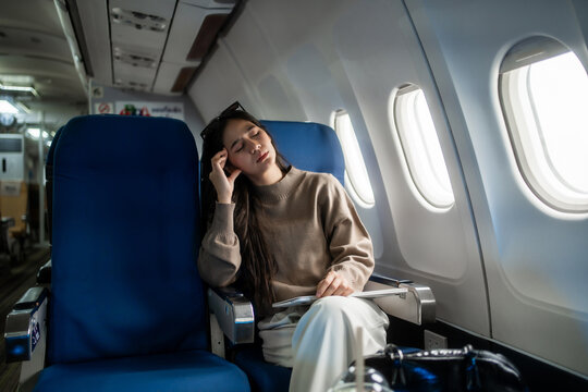 Tired young woman sitting by an airplane window, rubbing her forehead during a flight. Travel stress, fatigue, jet lag and discomfort inside a modern passenger cabin.