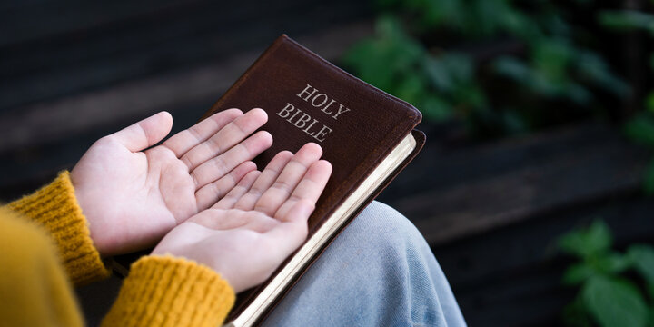 Woman holding hands open in worship and prayer over closed Holy Bible, seeking divine guidance and grace.