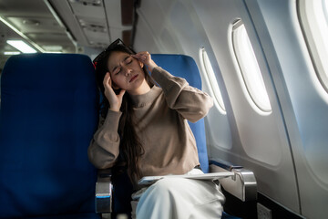Tired young woman sitting by an airplane window, rubbing her forehead during a flight. Travel stress, fatigue, jet lag and discomfort inside a modern passenger cabin. © Thitaporn