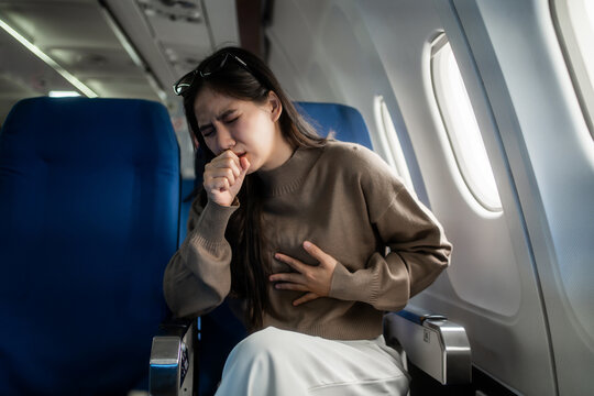 Young woman coughing and holding her chest while sitting by the window on an airplane, showing travel illness, discomfort, and in-flight health concern during a flight.
