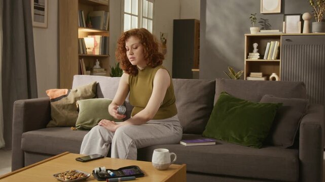 Caucasian young adult woman sitting on sofa, scanning continuous glucose monitor sensor on arm and checking blood sugar levels on smartphone. Perfect for diabetes care and digital health ads