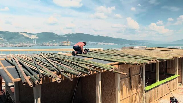 Unrecognizable shirtless builder constructing bamboo roof of beach canopy on seashore during hot summer day. Handheld panning wide shot with calm sea, port bay, and high mountains in background.