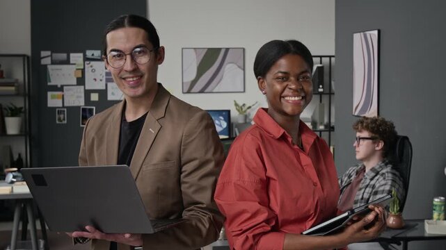 Waist-up portrait of Asian young man holding laptop and African American gen Z woman holding tablet, smiling at camera in office. Clip for tech startup marketing and corporate promos