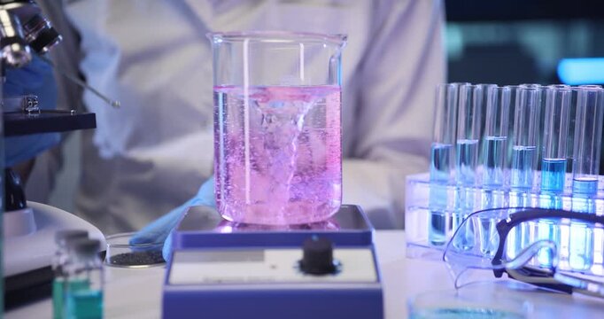 Scientist heats pinky solution on magnetic stirrer in lab. Gloved hand monitors bubbling mixture preparing reaction for further testing slow motion