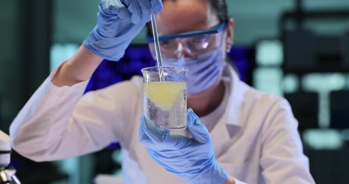 Woman scientist mixes yellow liquid inside clear beaker. Gloved hand lowers pipette stiring cloudy reagent during active lab reaction slow motion
