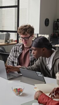 Vertical video of multiethnic young programmers collaborating on laptop and tablet in office. Footage for IT recruitment and software engineering promotional videos