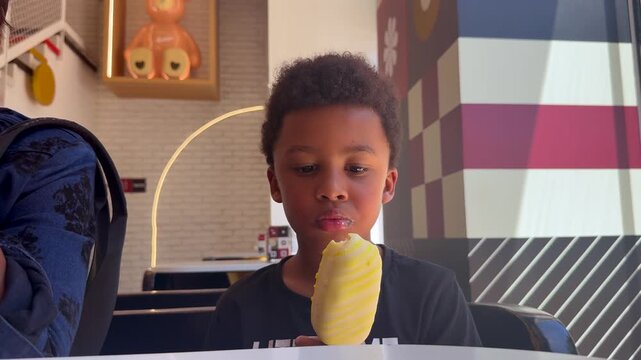 Close-up view of a 6-year-old biracial black toddler enjoying a vanilla ice cream sitting next to his mother inside an ice cream shop