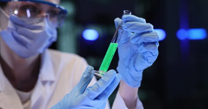 Woman scientist holds test tube with green liquid in lab. Gloved hand rotates sample inspecting contents during careful research check slow motion