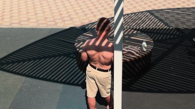 Shirtless muscular man leaning on white pillar looking at smartphone at outdoor beach gym. Slatted canopy casts striped shadows on his body. Handheld tracking observer shot moving right to left.