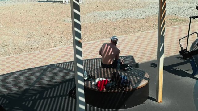 Unrecognizable shirtless man in cap sitting on round bench talking on smartphone. Slatted canopy casts geometric striped shadow patterns on his back. Handheld walking observer perspective.