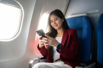 Young woman sitting by an airplane window, smiling while using her smartphone during a flight. Bright natural light and a calm travel mood make it ideal for business and leisure themes. © Thitaporn