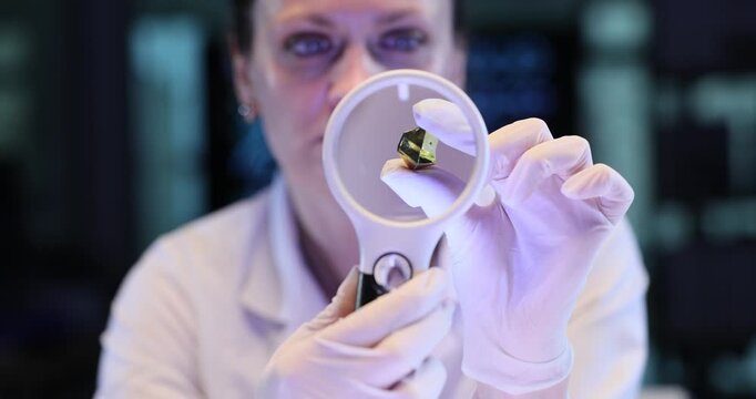 Woman scientist examines gold crystal through magnifier in lab. Gloved hand turns gemstone checking facets under focused blue light slow motion