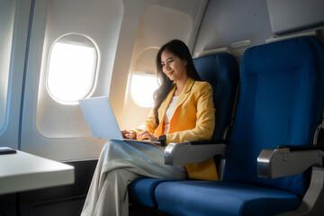 Young woman using a laptop while sitting by the window on an airplane, enjoying a calm flight. Ideal for travel, business, remote work, tourism, and modern lifestyle themes. © Thitaporn