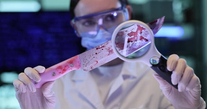 Female criminalist studies bloody knife with magnifying glass in lab. Forensic scientist woman analyzes weapon of crime in police laboratory