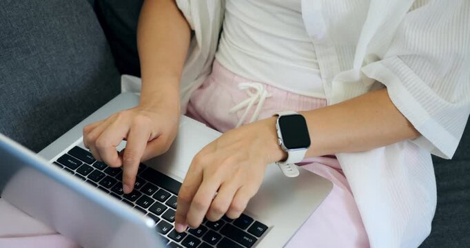 Woman uses wearable. Woman in living room focused on laptop with smartwatch alerts visible. Casual setting where woman types on her laptop while wearing smartwatch displaying notifications