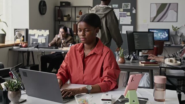 African American young woman typing on laptop and using smartphone in busy open-plan office. Footage for corporate training videos and technology marketing campaigns