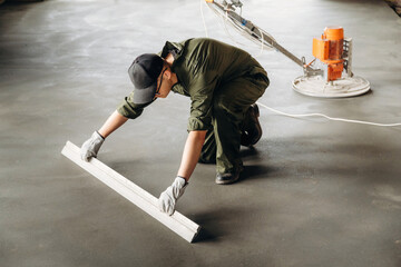 Obraz premium Male construction worker in green coveralls uses a leveling tool on freshly poured concrete floor with a power trowel visible in the background