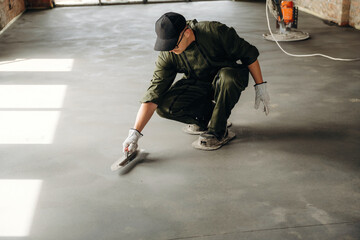 Obraz premium Male construction worker in green coveralls kneeling on a concrete floor applying finishing touches with a trowel in an industrial setting with natural light