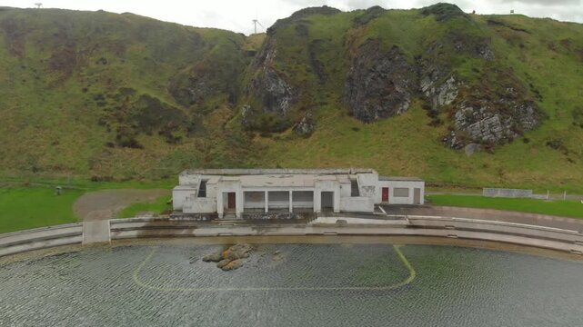 Drone retreats from derelict Tarlair lido, cliffs and wind turbines.