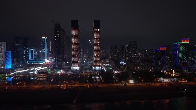 Lateral drone night shot of Batumi seafront capturing illuminated modern buildings, vibrant skyline, and reflections along the Black Sea coastline.