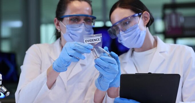 Female technicians in robes observe glass vial in plastic bag in lab. Forensic scientists analyze crime evidence in police laboratory slow motion