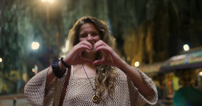 Woman makes heart shape with hands at Batu Caves in Malaysia during evening time with colorful lights in background