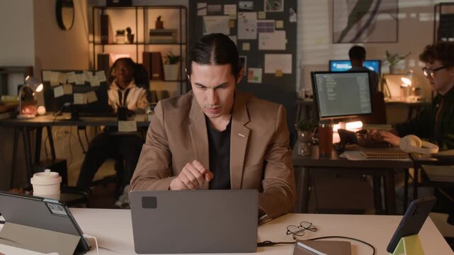 Young Asian man working on laptop in office at night, celebrating success with multiethnic colleagues. Clip for tech startup promotions and corporate teamwork videos