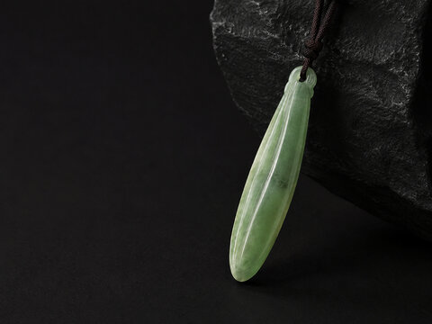 A slender green jade pendant shaped like a silique on a dark background.