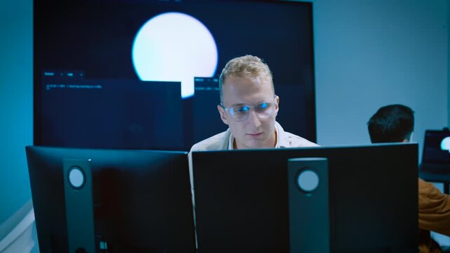 Focused young male IT specialist typing code and looking at multiple computer monitors while developing AI software system in tech office