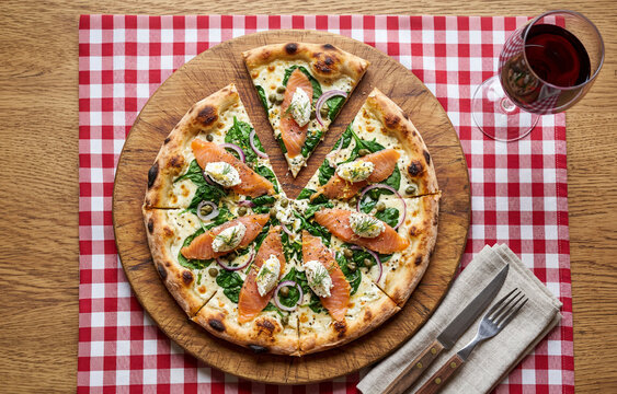 Tasty pizza with salmon, cream cheese and baby  spinach on wooden board in  a restaurant table setting with glass of red wine