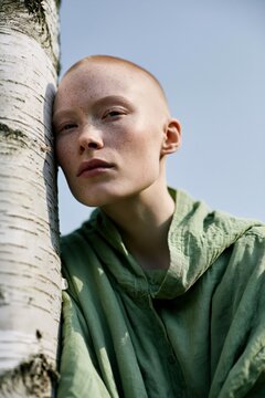 Young woman with shaved head leaning on birch tree in green linen hoodie natural light minimalist portrait banner