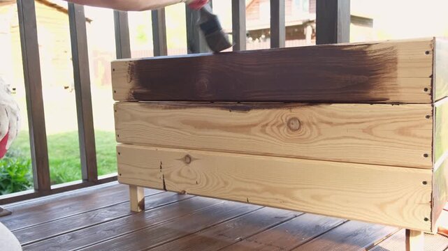 Brown oil for protection of wood by staining. Painting fence for protecting and pests. Woodworker applying finish to pine board box. Repairman varnishing with brush wooden fencing wall plank surface.