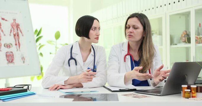 Pair of women doctors discusses patient records at clinic desk. Specialists gesture with hands explaining treatment at teamwork meeting slow motion