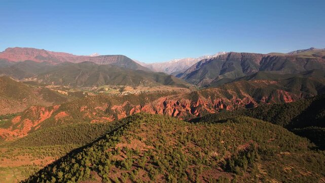 Ourika Valley, Al Haouz Province, Marrakesh Safi, Morocco, featuring red sedimentary ridges, juniper woodland and snow capped High Atlas peaks including the Toubkal massif, drone forward flight shot.