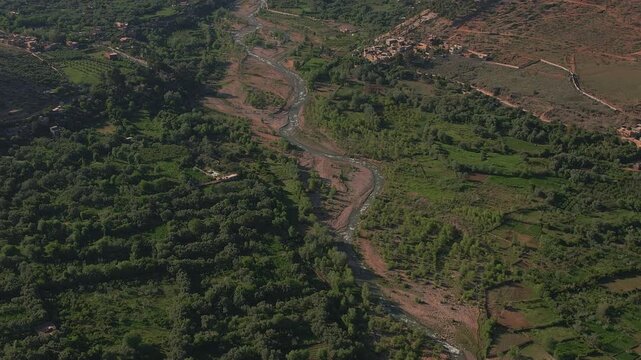 Ourika River flowing through the Ourika Valley, Al Haouz Province, Marrakesh Safi, Morocco, featuring braided riverbeds, oasis vegetation and irrigated agricultural terrain, zoom out ascent drone shot
