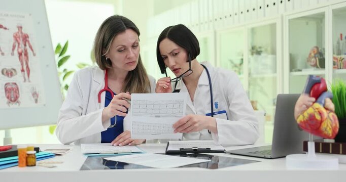 Female doctors review medical chart at clinic desk. Specialists point at printed data discussing findings during focused consultation slow motion