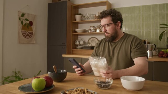 Medium shot of adult man with diabetes sitting at kitchen table, checking food package nutrition facts on smartphone. Footage perfect for healthcare apps, diabetes tracking, wellness marketing