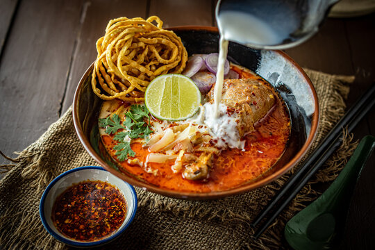 Action shot of pouring soup into a traditional bowl of Northern Thai Khao Soi chicken curry noodle with crispy egg noodles and lime, served on a dark rustic wooden background table setting.