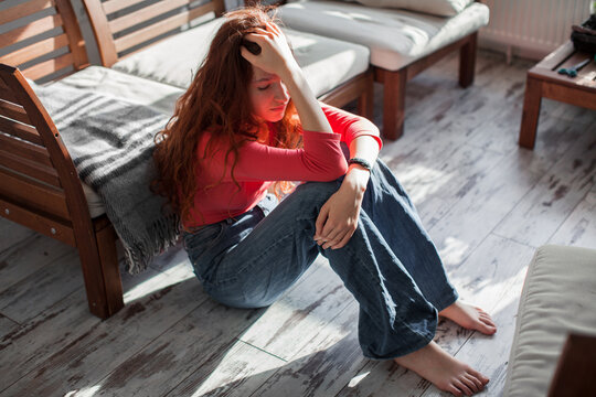Sad teenage girl sitting on the floor in a room. Depressed woman holding head with hand feeling lonely and upset. Mental health crisis, psychological trauma and stress concept.
