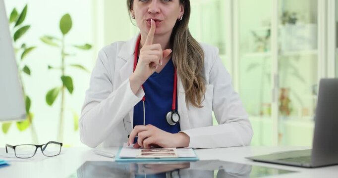 Woman doctor makes silence gesture at clinic desk. Specialist places finger near lips requesting quiet during focused medical consultation slow motion
