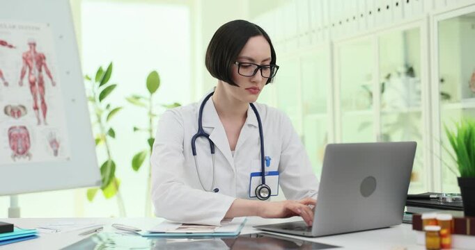 Woman doctor types on laptop at bright clinic desk. Specialist reviews digital records working through patient notes with focused face slow motion