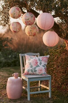 Weathered grey wooden chair with floral pillow and pink lanterns hanging from a tree in a garden corner during sunset, creating a serene outdoor setting