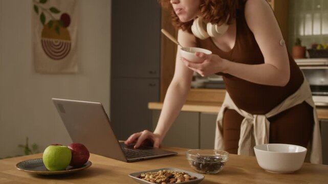 Cropped shot of pregnant Caucasian young adult woman with CGM sensor on arm typing on laptop and holding bowl in kitchen. Clip is perfect for gestational diabetes marketing