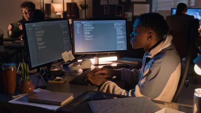 Young African American programmer typing code on dual computer monitors during night shift in office. Video for IT recruitment, software development, and tech education marketing