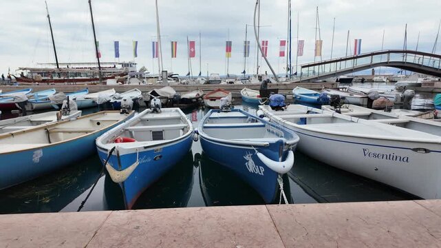 Bardolino, Italy - May 14, 2026 - Boats docked in a harbor with flags and a pedestrian bridge overhead