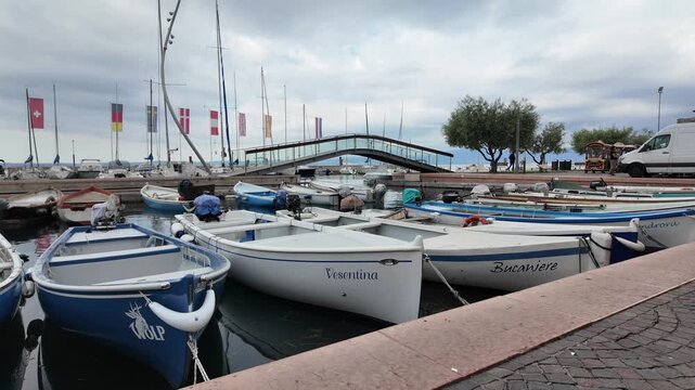 Bardolino, Italy - May 14, 2026 - Boats docked in a harbor with flags and a pedestrian bridge overhead