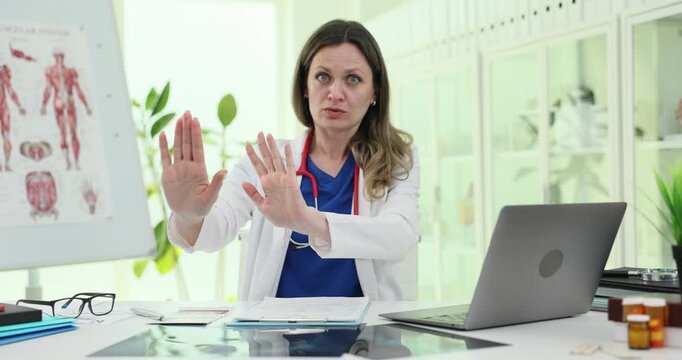 Woman doctor rejects request at bright clinic desk. Specialist raises palms giving firm No gesture during calm medical consultation slow motion