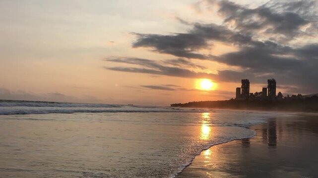 Ocean waves reach a sandy shoreline during sunset, creating golden reflections on wet sand beneath a colorful sky with dramatic clouds and coastal scenery.