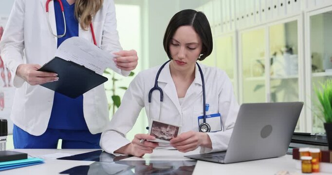 Woman colleague brings documents to doctor at clinic desk. Seated specialist looks up reviewing scan images during clinic consultation slow motion