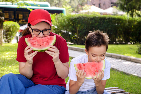 Mother and son enjoying juicy watermelon slices on a park bench during a sunny summer day. Healthy family snack, outdoor bonding, childhood happiness, and seasonal lifestyle concept